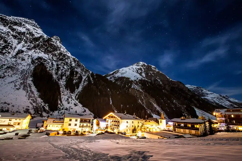 Snowy Plangeross with illuminated houses and church tower under the starry sky, high mountains behind