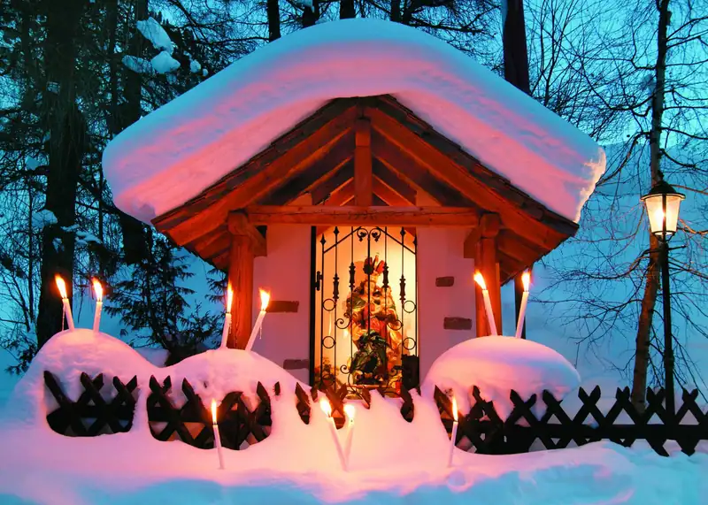Snow-covered wayside chapel with a statue of Mary, candles burning in the snow in front