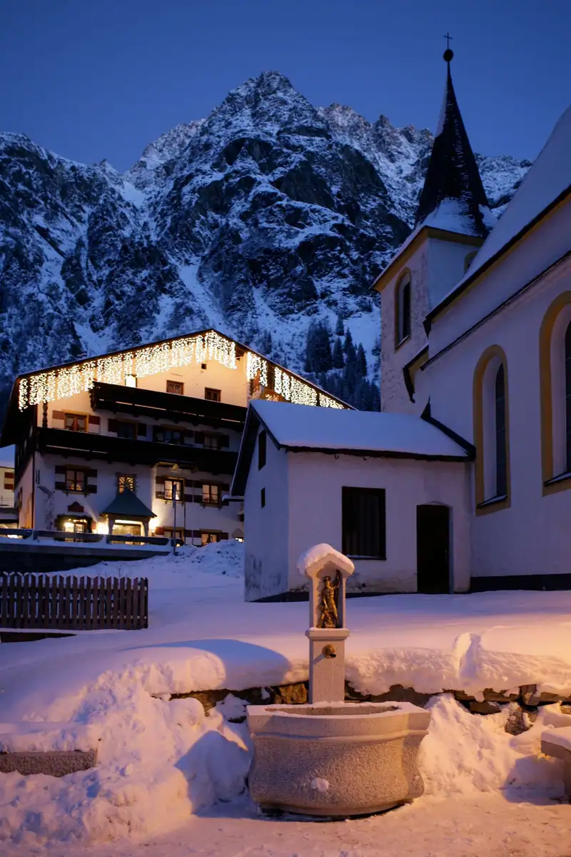 Snow-covered village fountain in front of the church and a house with fairy lights, a rocky peak behind