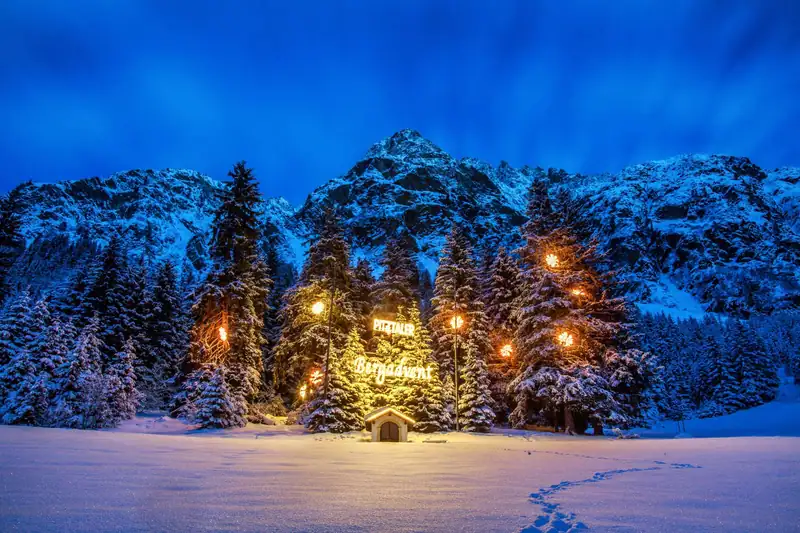 Illuminated lettering “Pitztaler Bergadvent” in snowy spruces in front of a mountain peak at blue hour