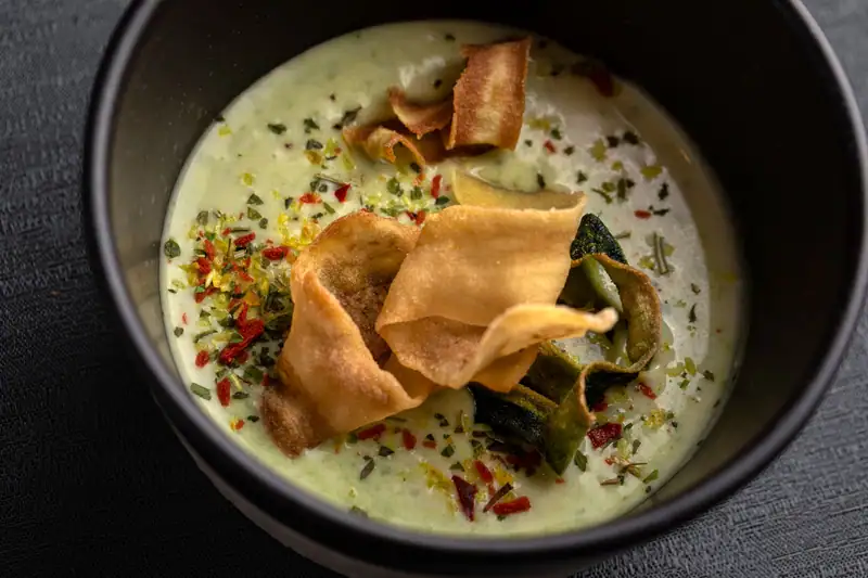 Creamy herb soup with crispy vegetable chips and colourful spice flakes in a dark bowl