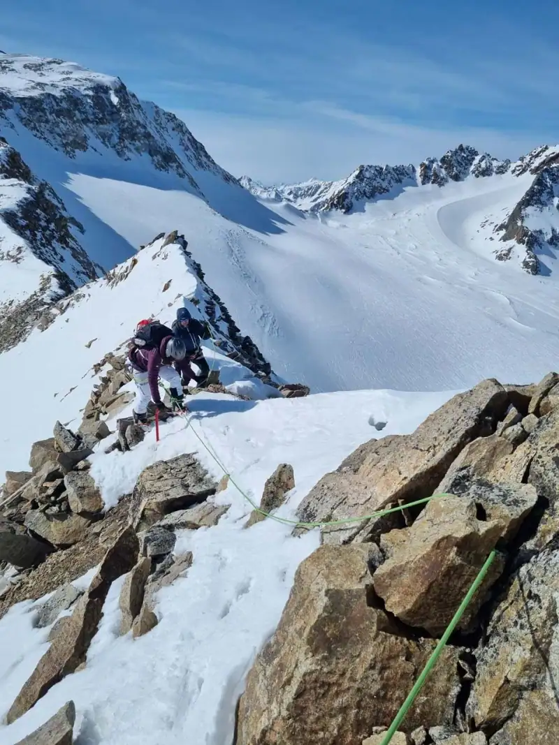 Two mountaineers roped together on the rocky, snowy summit ridge, a wide glacier behind