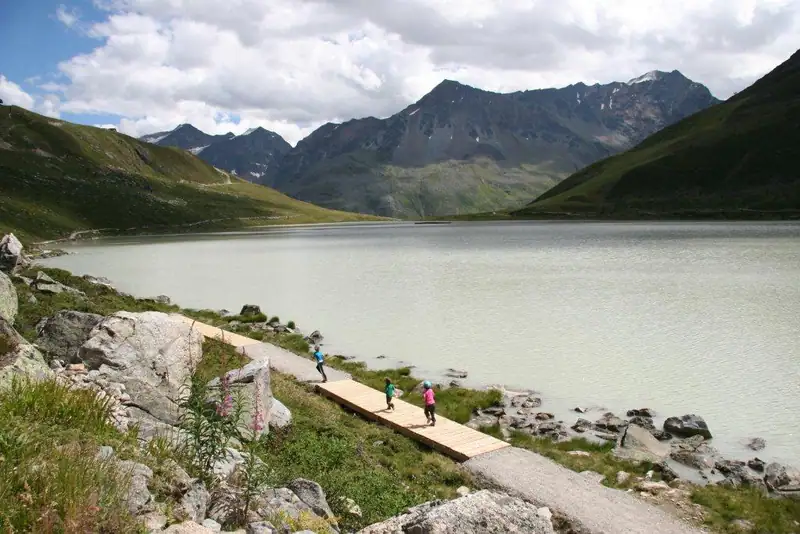 Children running over a wooden boardwalk on the shore of the turquoise-grey Rifflsee, mountains behind