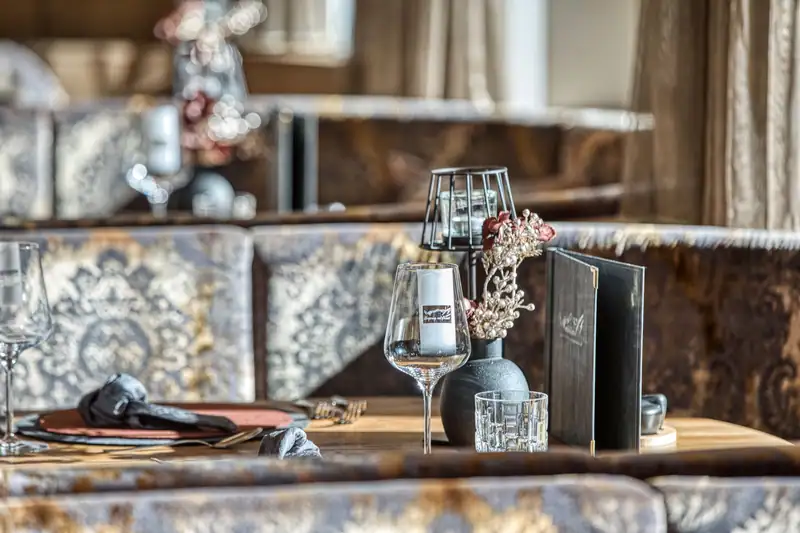 Gedeckter Tisch mit Weinglas, Windlicht, Blumenvase und Speisekarte vor gold-grauen Polsterbänken