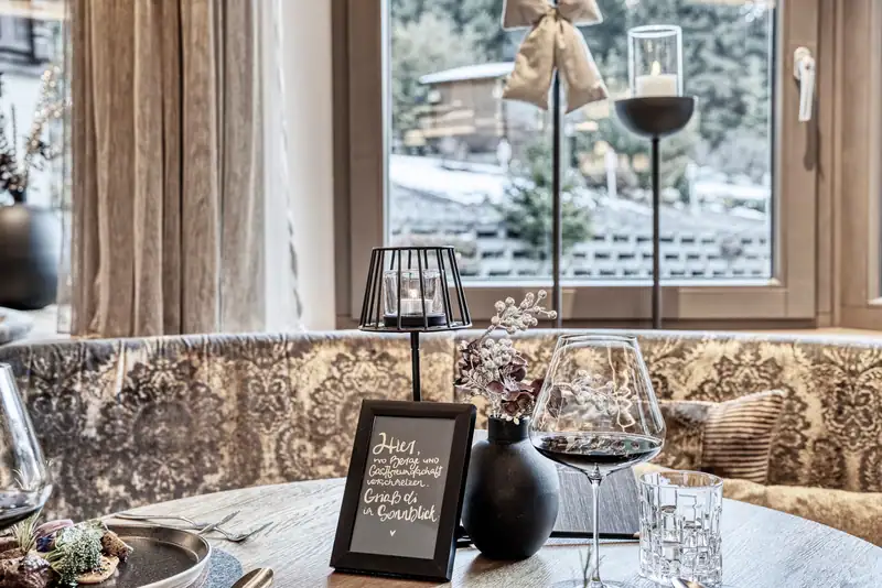 Table by the window with lantern, red wine and a sign “Where mountains and hospitality merge. Griaß di at Sonnblick”