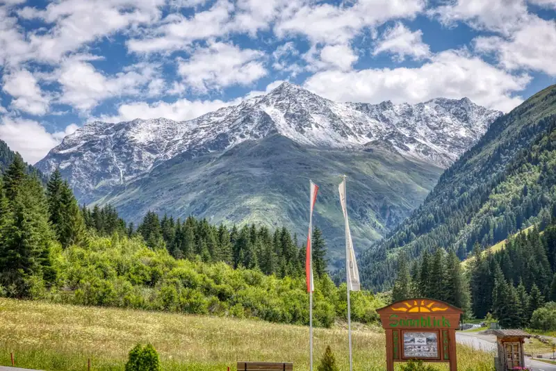 Wooden Sonnblick sign and flags at the entrance, a snow-capped peak behind in summer