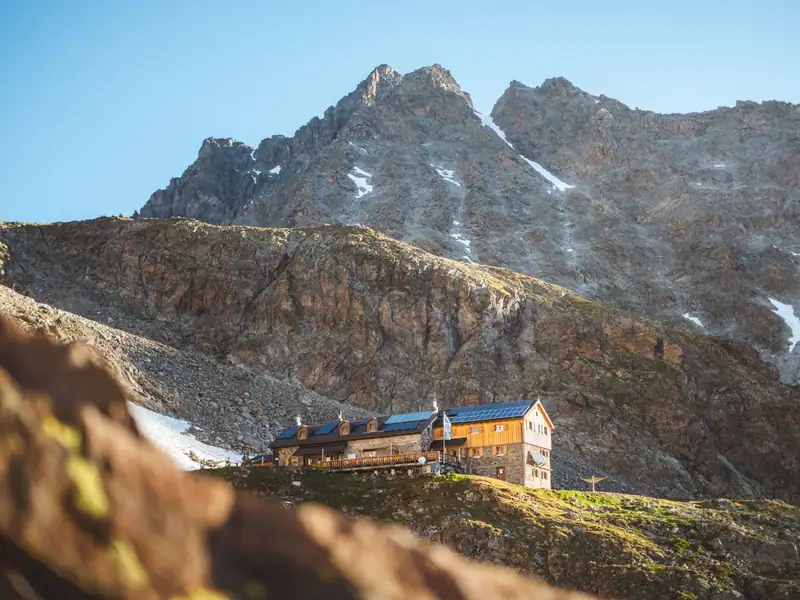 Stone and wood mountain hut with solar roof beneath jagged rocky peaks