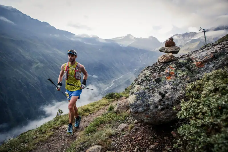 Trail runner with poles on a mountain path above a cloud-covered valley