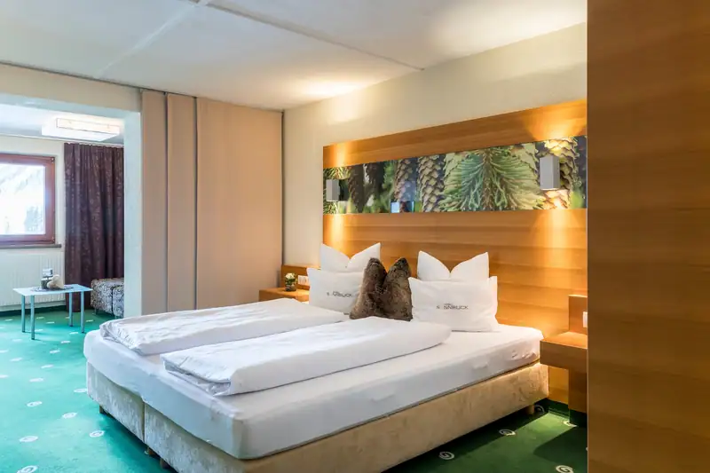 Box-spring bed in front of a wooden wall with pine-cone motif, the living area with bench seat and window on the left